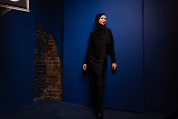 Fashion muslim woman in black hijab is posing on blue background in studio. Professional caucasian...
