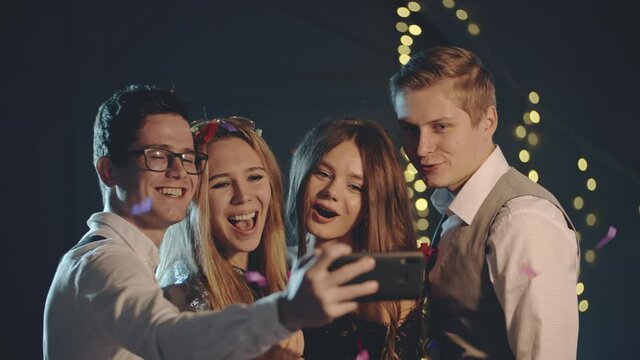 Smiling multiracial friends throw confetti, posing and taking selfie on smartphone or mobile phone. Happy guys having fun and taking photo for memory using modern technology at Christmas party.