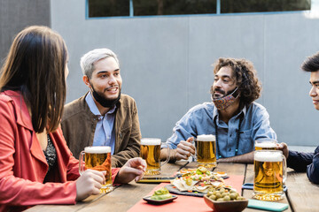 Multiracial people having fun drinking beer at brewery bar outdoor while wearing safety masks - Focus on left guy face