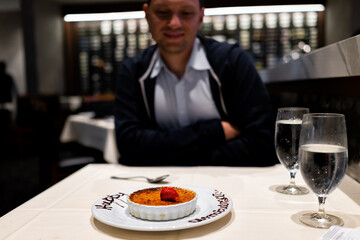Young happy man sitting inside restaurant for dinner looking at food on date with French creme brulee dessert with piped happy anniversary sign