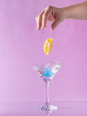 Woman hand throwing lemon into martini glass full with alcohol drink with splashes, against pastel...