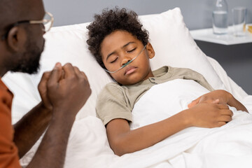 Close-up of African little boy with tube in his nose for breathing sleeping in bed while father...