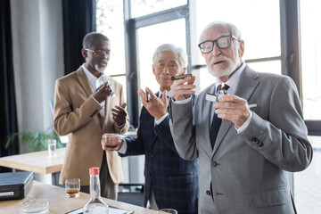 senior respectable businessman holding lighter and cigar near blurred interracial colleagues with glasses of whiskey