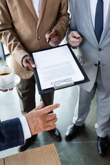cropped view of senior businessman with cup of coffee near interracial colleagues with contract on clipboard