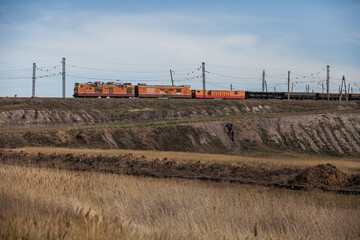 Train with orange locomotive and wagon in steppe. Platform cars for coal and ore transportation. Yellow grass, brown ground. Electric towers, wires, blue sky with cloud. Sunset view.