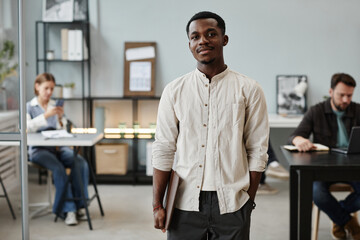 Naklejka na ściany i meble Portrait of African young businessman looking at camera while standing at office with his colleagues in the background