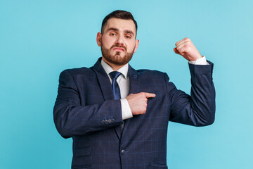 Egoistic self confident man wearing official style suit pointing finger at arm biceps, showing his strength and independent, leadership concept. Indoor studio shot isolated on blue background.