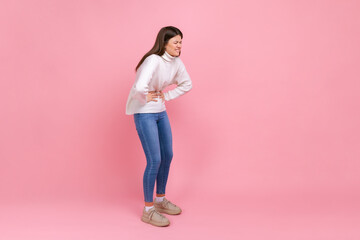 Full length portrait of unhealthy woman suffering strong stomachache, terrible pain during period, wearing white casual style sweater. Indoor studio shot isolated on pink background.