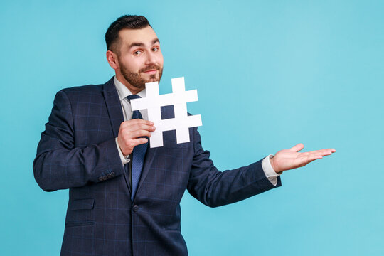 Man wearing official style suit, holding white hashtag and presenting at copy space on her palm, empty place for idea presentation, product advertising. Indoor studio shot isolated on blue background.