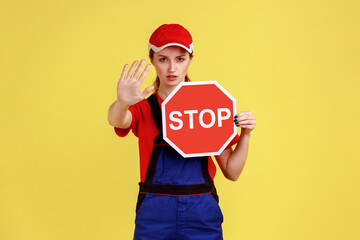 Portrait of worker woman showing stop gesture and holding red stop road sign, prohibitions and restrictions, wearing overalls and red cap. Indoor studio shot isolated on yellow background.