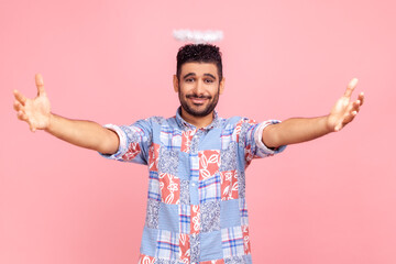 Come into my arms! Portrait of attractive angelic bearded guy with nimb over head in blue casual style shirt, spreading arms, invites hugging. Indoor studio shot isolated on pink background.