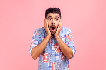 Portrait of astonished bearded man in blue shirt looking with big eyes, holding hands on face and...