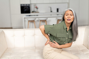 Gray-haired woman felt a sharp pain in loin sitting on the sofa at home, worried Asian lady holding...