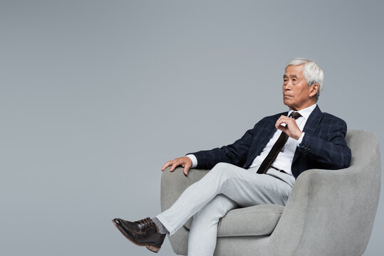 Senior And Confident Asian Businessman Looking Away While Sitting In Armchair Isolated On Grey