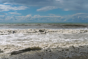 stormy waves of the Mediterranean sea run over the rocky summer beach against the backdrop of a blue cloudy sky