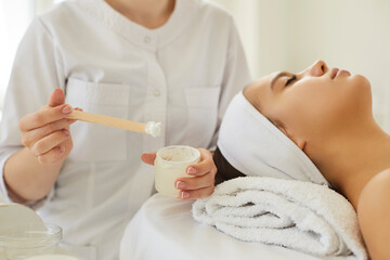 Obraz na płótnie Canvas Female cosmetologist stirs and prepares moisturizing cosmetic mask to apply it to client's skin. Close up of jar with white mask or cream in hands of beautician sitting near head of female client.