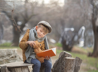 Fototapeta premium a boy in a yellow jacket sits on a bench with a toy bear and reads him a book in autumn. the concept of loneliness, the search for a friend.