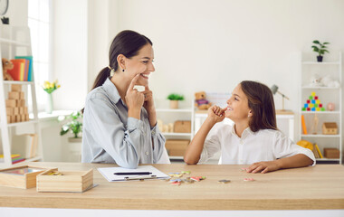 Female specialist working on kid's speech impediments. Smiling child together with professional language therapist sitting at desk in modern office, playing fun games, developing correct pronunciation