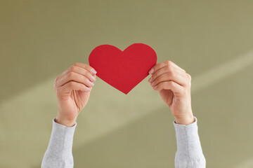 Close up of young woman's hands holding red paper heart as sign of blog likes, sympathy or approval symbol. Woman holding heart on green background. Concept of social networking, love or charity.