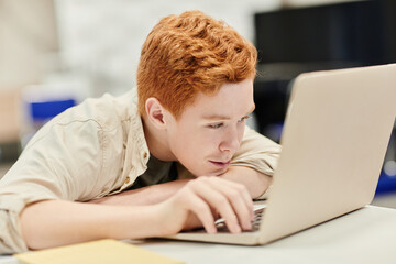 Portrait of red haired teenage boy laying head on desk while using laptop in school, copy space