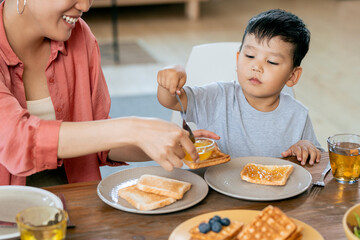 Happy Asian female helping her little son to make sandwich with honey or jam while having breakfast