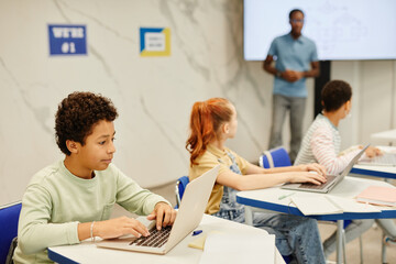 Side view portrait of teenge boy using laptop while studying in modern classroom with group of children, copy space