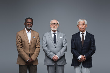 senior multiethnic business partners in suits looking at camera isolated on grey