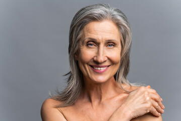 Stunning attractive elderly gray-haired woman look at the camera over grey background, charming mature lady gently touch her naked shoulder, has beautiful smile with white healthy teeth