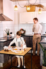 Happy young caucasian family, kid making baking cookies in modern kitchen while handsome american dad father in the background washing the dishes, at home. family time. single dad lifestyle