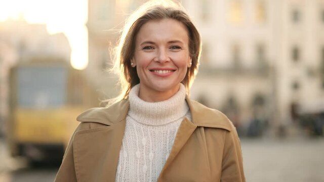 Lovely middle-aged blond woman with a beaming smile and looking at the camera. Happy adult woman smiling with teeth smile outdoors and stay on city street at sunset time