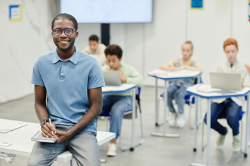 Obraz premium Portrait of young African-American teacher sitting on desk and smiling at camera with children in background, copy space