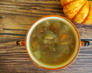 mushroom soup with Polish mushrooms, carrots, potatoes and onions in a brown plate with handles stands on a brown wooden background next to a small pumpkin. top view