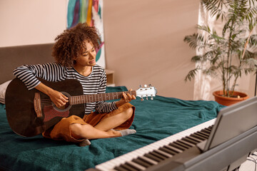 Female musician sitting on bed near synthesizer and playing melody on acoustic musical instrument