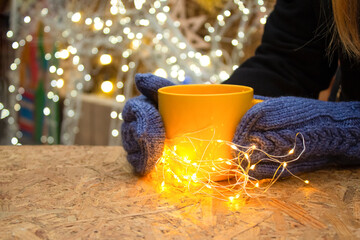 Woman's hands holding yellow mug with hot tea or coffee with illumination lights around. Christmas market with illuminating background