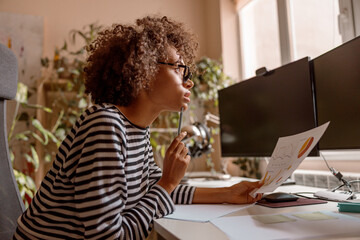 Female freelancer sitting at the table and holding sheet of paper with graphs while pondering about best solutions for project