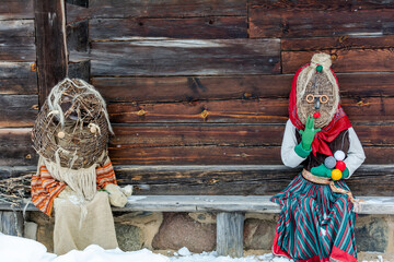 Dolls in Meteņi mumming masks and costumes sitting in a bench near an old wooden house. Meteni is...