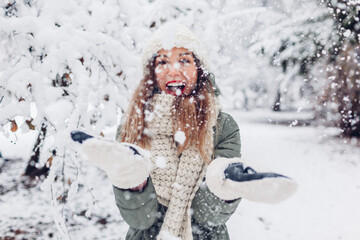 Happy young woman playing with snow in snowy winter park wearing warm knitted clothes and having fun.