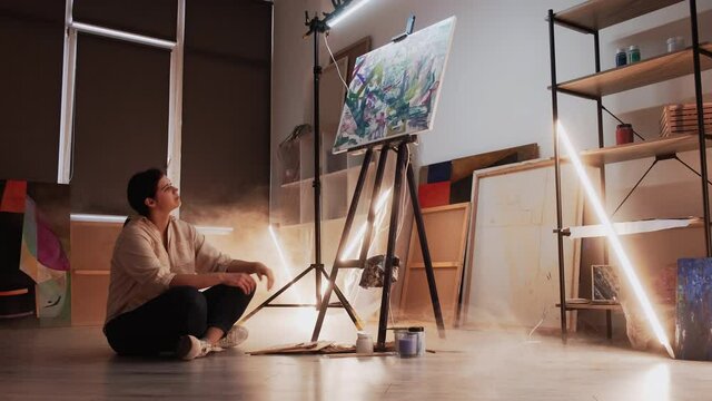 Finish artwork. Enjoy result. Art studio. Happy woman sitting floor looking on abstract picture in dark light room interior.