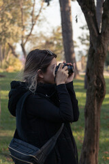 Female photographer, taking pictures with her digital camera of forest landscape at sunset. A girl in black is taking photograph in forest. Green background. Unrecognizable person. 