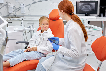 Professional caucasian redhead female dentist communicating with cute little child patient while checking teeth during dental procedure at modern bright dentist's office. Side view. Health care