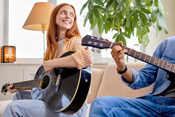 Happy woman sitting comfy sofa playing guitar learning spend time with man together stay home quarantine safety modern interior living room indoors, redhead lady and handsoem guy in casual wear