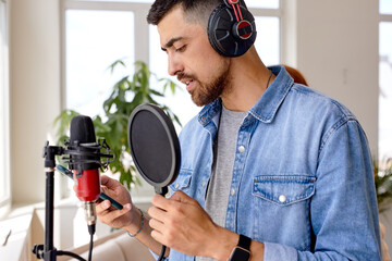 music, show business, people and voice concept.handsome male singer with headphones and microphone singing song at sound recording studio, holding smartphone in hands, side view portrait