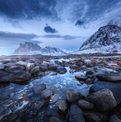 Seashore with stones and blurred water, against snowy mountains and blue sky with clouds at dusk. Uttakleiv beach in Lofoten islands, Norway. Winter landscape with sea, waves, rocks at night. Nature