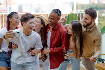 Young cheerful people having barbecue party on terrace by home, make selfie together, take photo on smartphone, having fun and talk. Multi-ethnic Group of friends having party, side view portrait