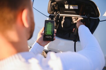Man Charging Electric Vehicle With Cable Looking At App On Mobile Phone