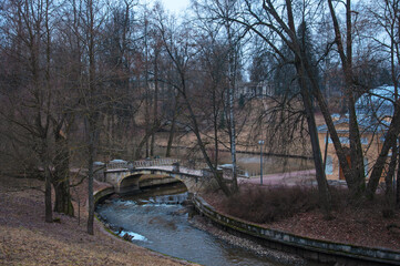 River and old beautiful stone bridge in city park with big old trees in late autumn