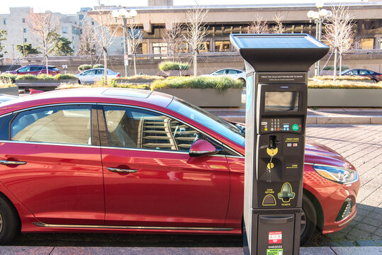Parking machine with a solar battery on a city street against the backdrop of a parked red car