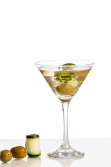 a martini glass with a drink decorated with olive and cucumber on a reflective surface, isolated on white background.