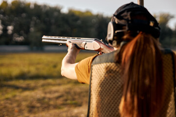 Rear view redhead caucasian woman on tactical gun training classes. Woman with weapon. Outdoor Shooting Range. Side view on beautiful lady with rifle machine gun in the forest, slim female is hunting - obrazy, fototapety, plakaty