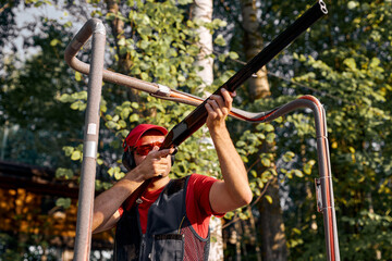 After getting shooting instructions, young confident focused man concentrated at aiming rifle at side, fires at target in outdoor range, wearing goggles, cap and headset equipment outfit